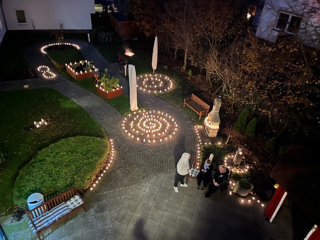 Beleuchteter Garten in der Abenddämmerung mit dekorativen Lichtern entlang der Wege und um runde Blumenbeete. Zwei Personen gehen durch die Anlage. Die Szene wirkt festlich, stimmungsvoll und liebevoll gestaltet.
